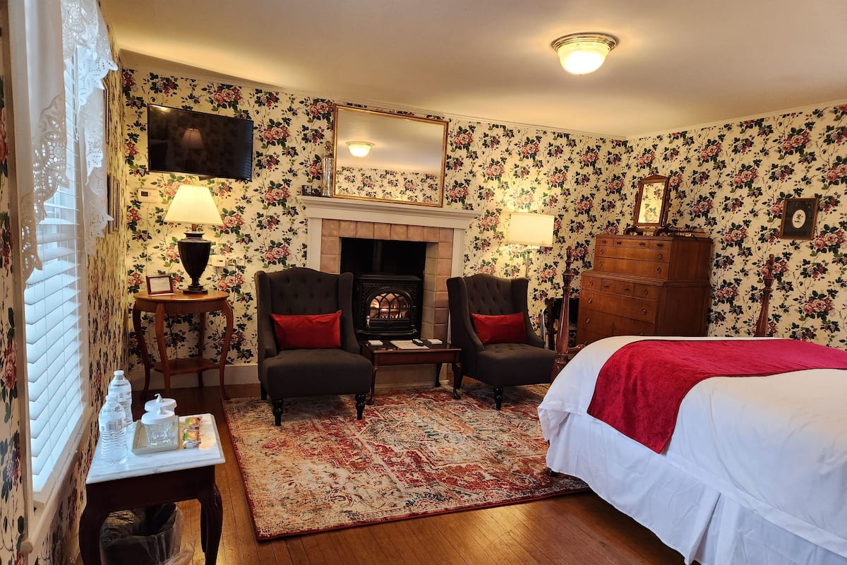 A hotel room with floral wallpaper, a fireplace, two armchairs, a bed, and a wooden dresser.