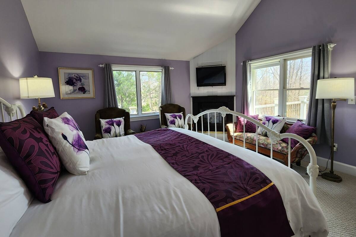 A bedroom with a large bed, two armchairs, a fireplace, and a TV on the wall.