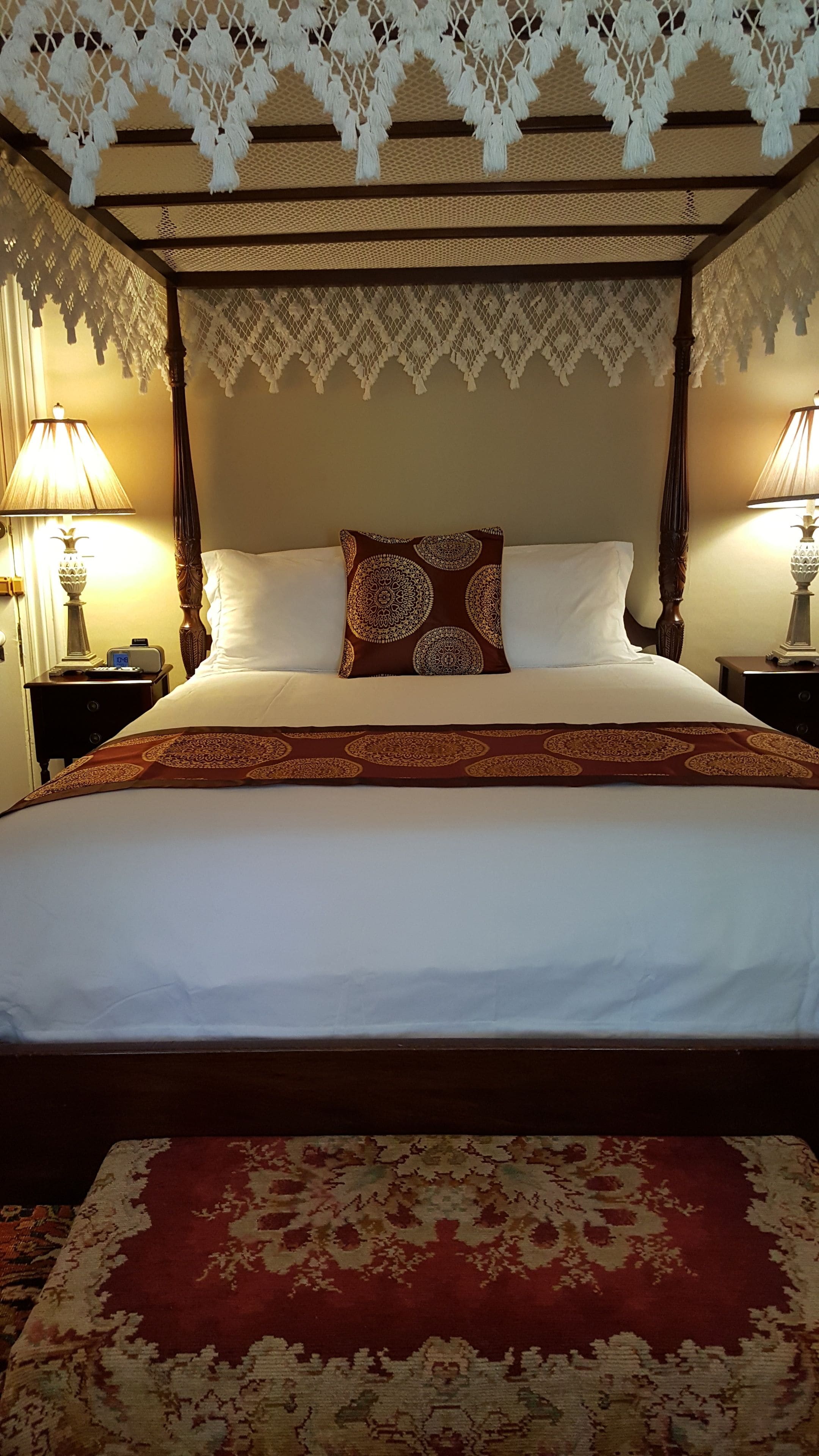 Una cama con dosel con un cojín estampado en blanco y marrón, una colcha rojo y beige, y una alfombra rojo y beige.