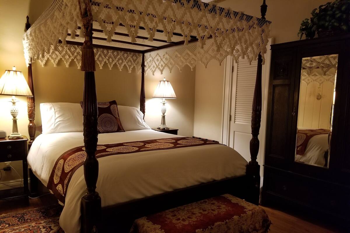 A four-poster bed with a canopy, two bedside lamps, a large mirror, and a patterned rug.