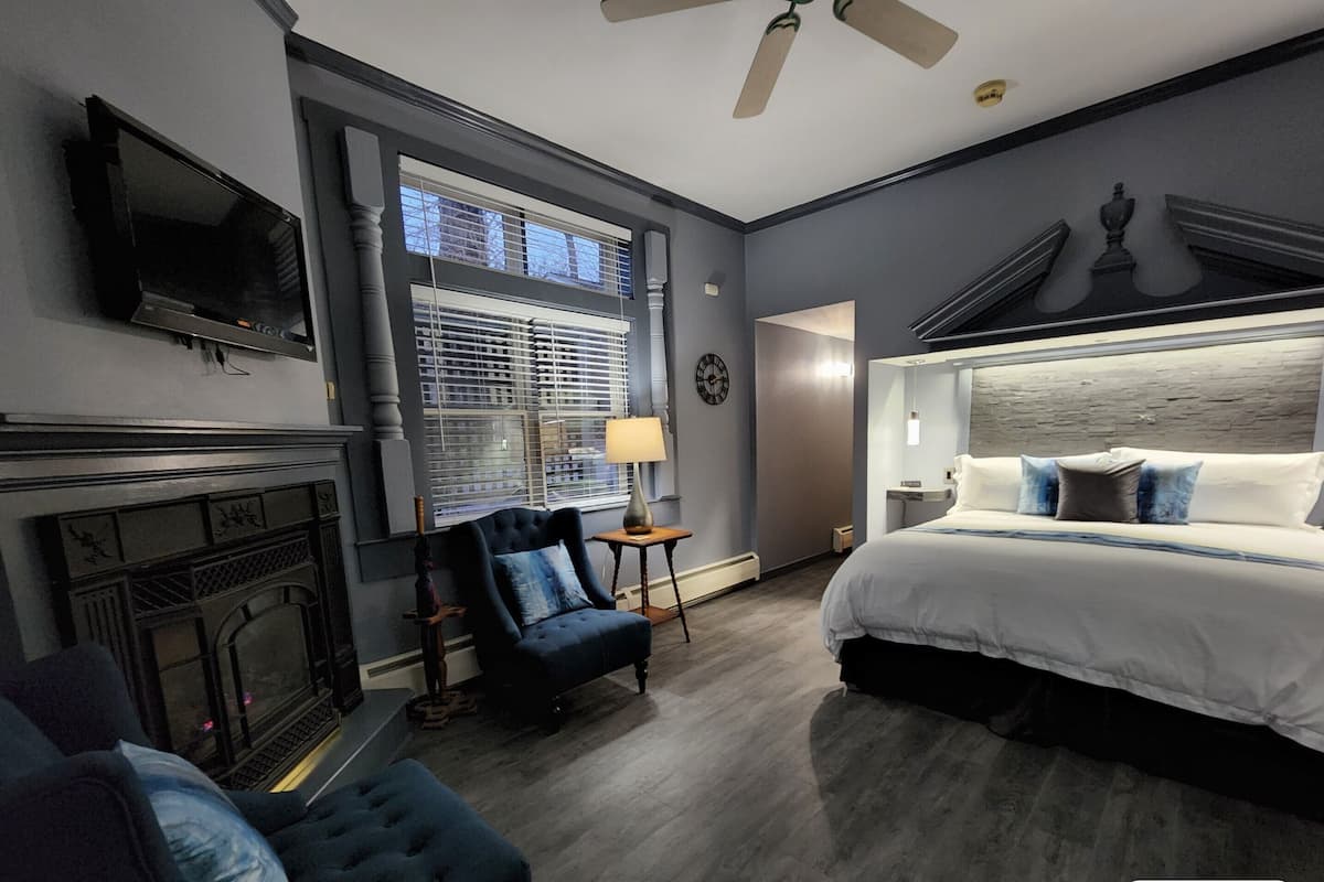 A modern hotel room with a fireplace, a large bed with a headboard, a TV mounted on the wall, and a ceiling fan.