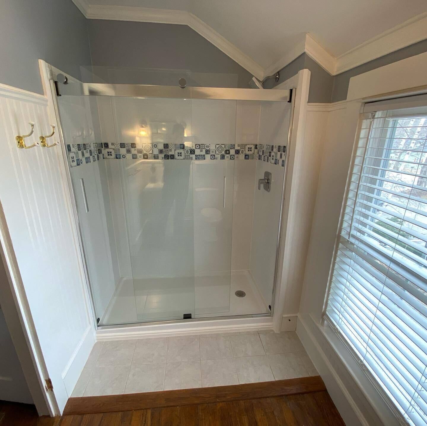 A glass-enclosed shower room with a tiled floor and a window with blinds.