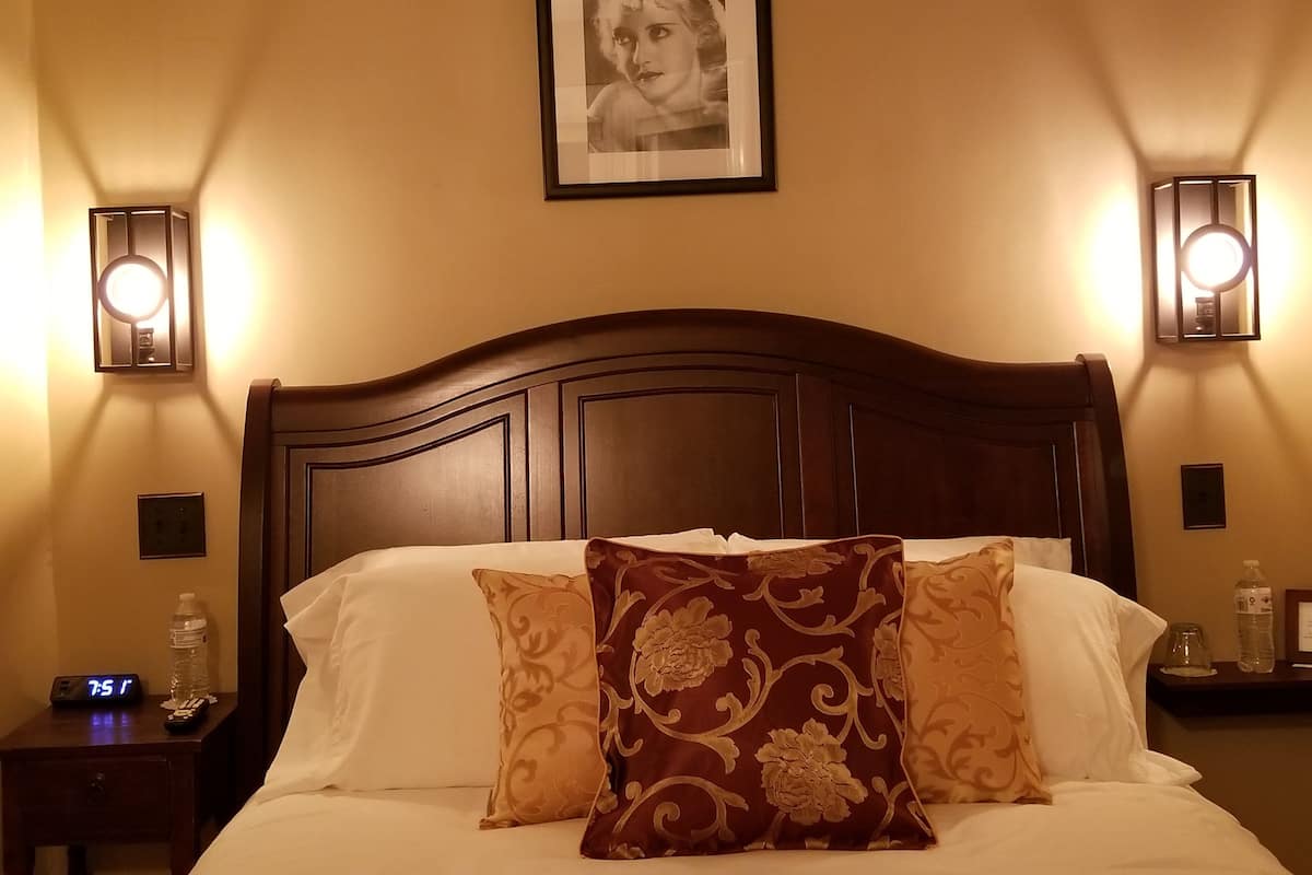 A hotel room with a wooden headboard, two bedside lamps, a framed picture on the wall, a digital clock, and a nightstand with a water bottle and remote control.