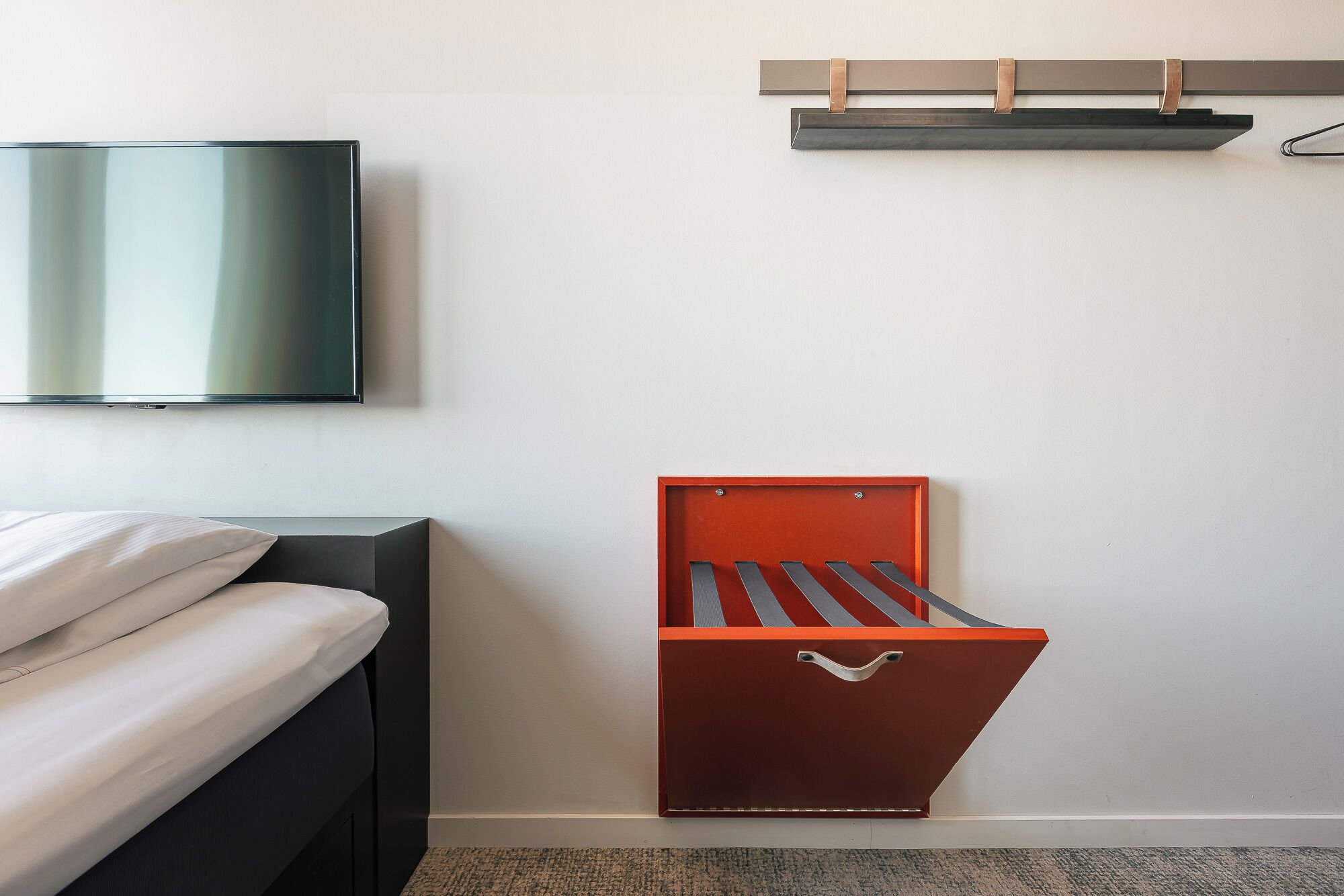 Une chambre à coucher avec un lit, un téléviseur fixé au mur et un meuble de rangement rouge doté d’une poignée.
