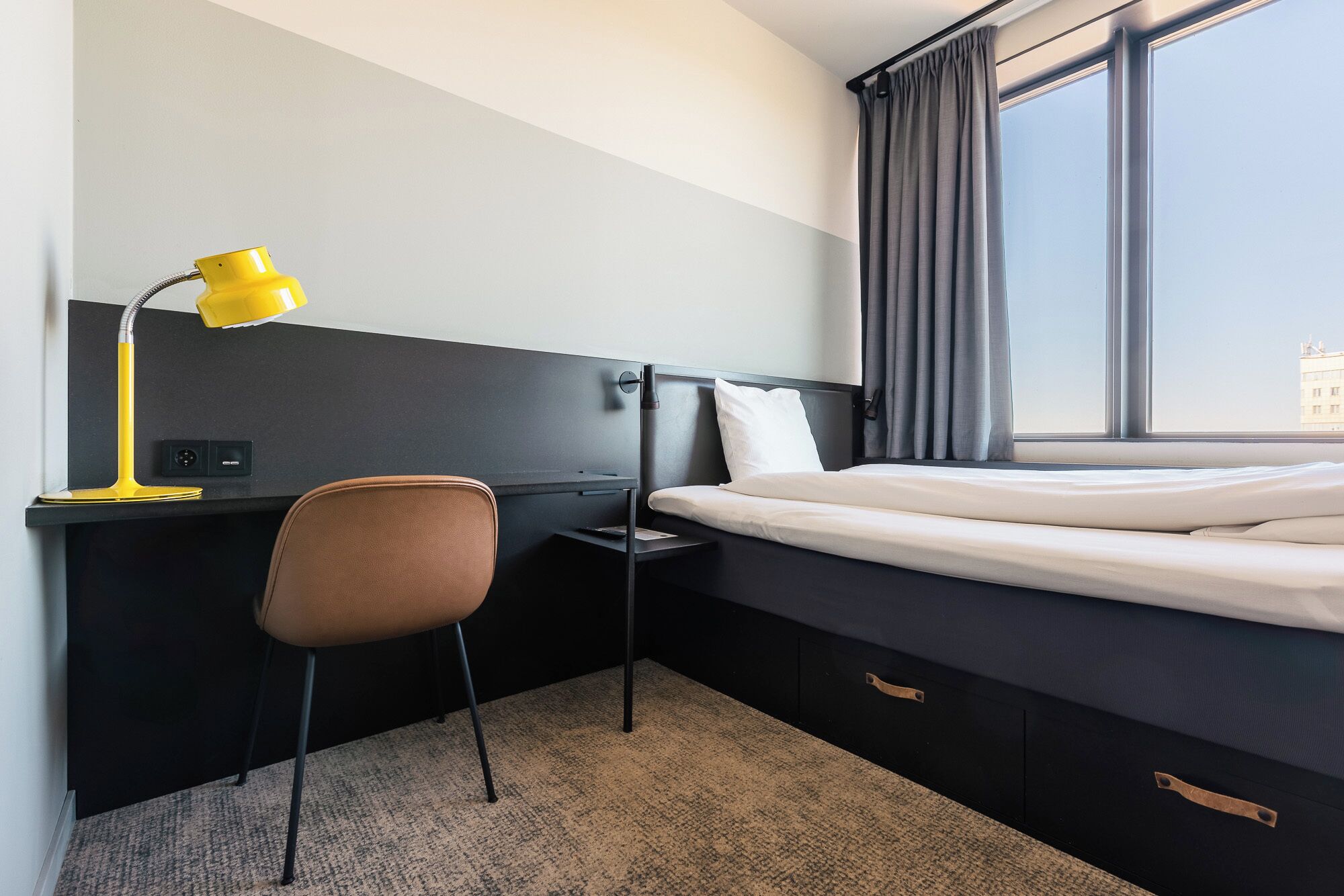 Habitación de hotel con cama, escritorio, silla, lámpara de escritorio y ventana con cortinas.