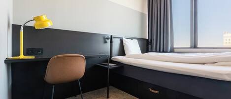 A hotel room with a bed, a desk with a chair, a desk lamp, and a window with curtains.