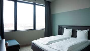 A hotel room with a large window, a bed with white linens, a dark headboard, and a small table.