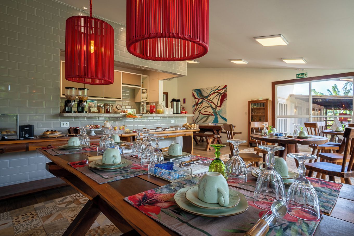 Ein Restaurant mit einem langen Esstisch, der für eine Mahlzeit gedeckt ist, einer Küche und Blick ins Freie.