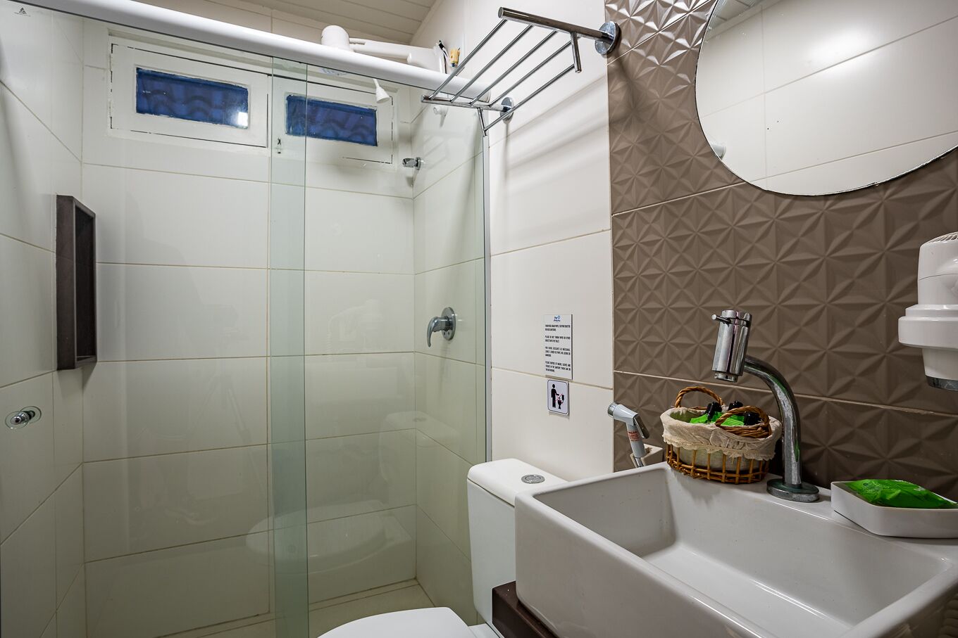 Un baño moderno con ducha de vidrio, un lavamanos blanco y un cesto de artículos de aseo, y una pared color beige con diseño.