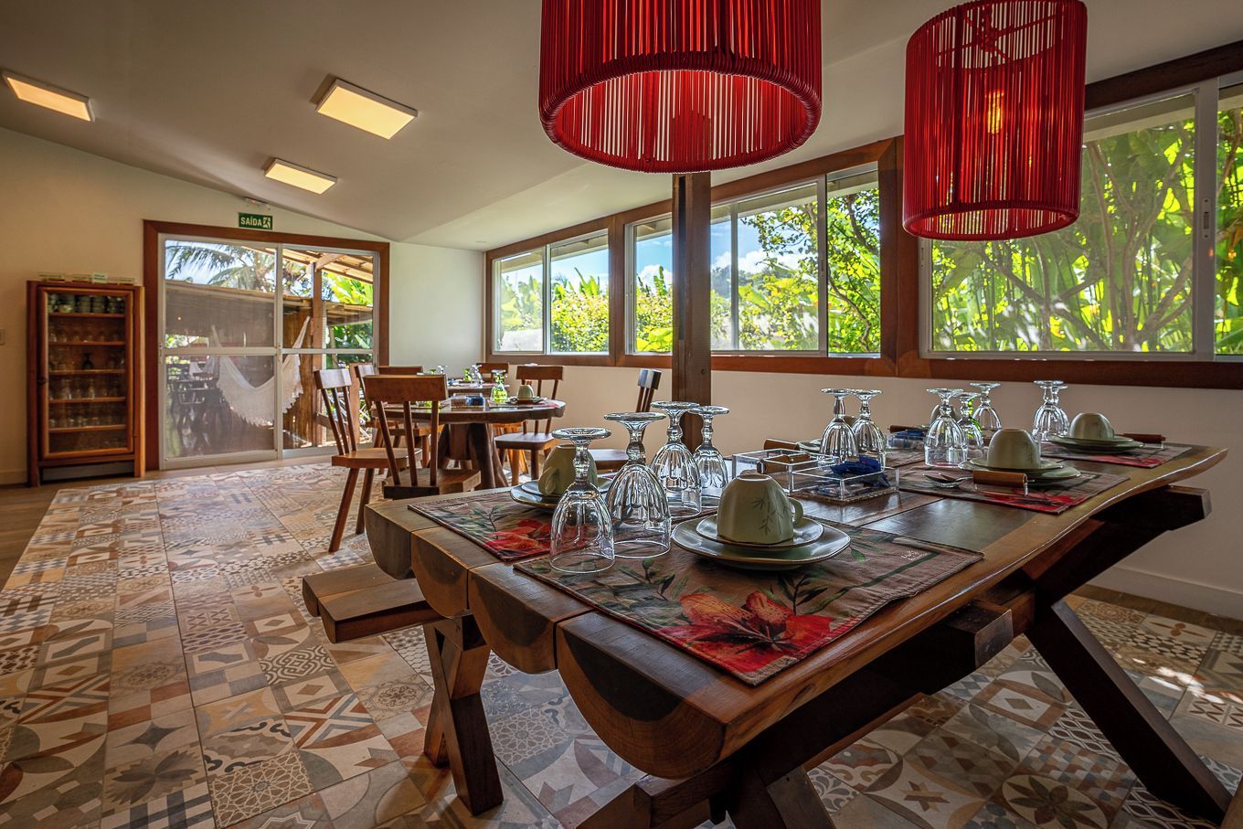 A dining area with a long table set for a meal, large windows with a view of greenery, and a wooden bookshelf in the background.