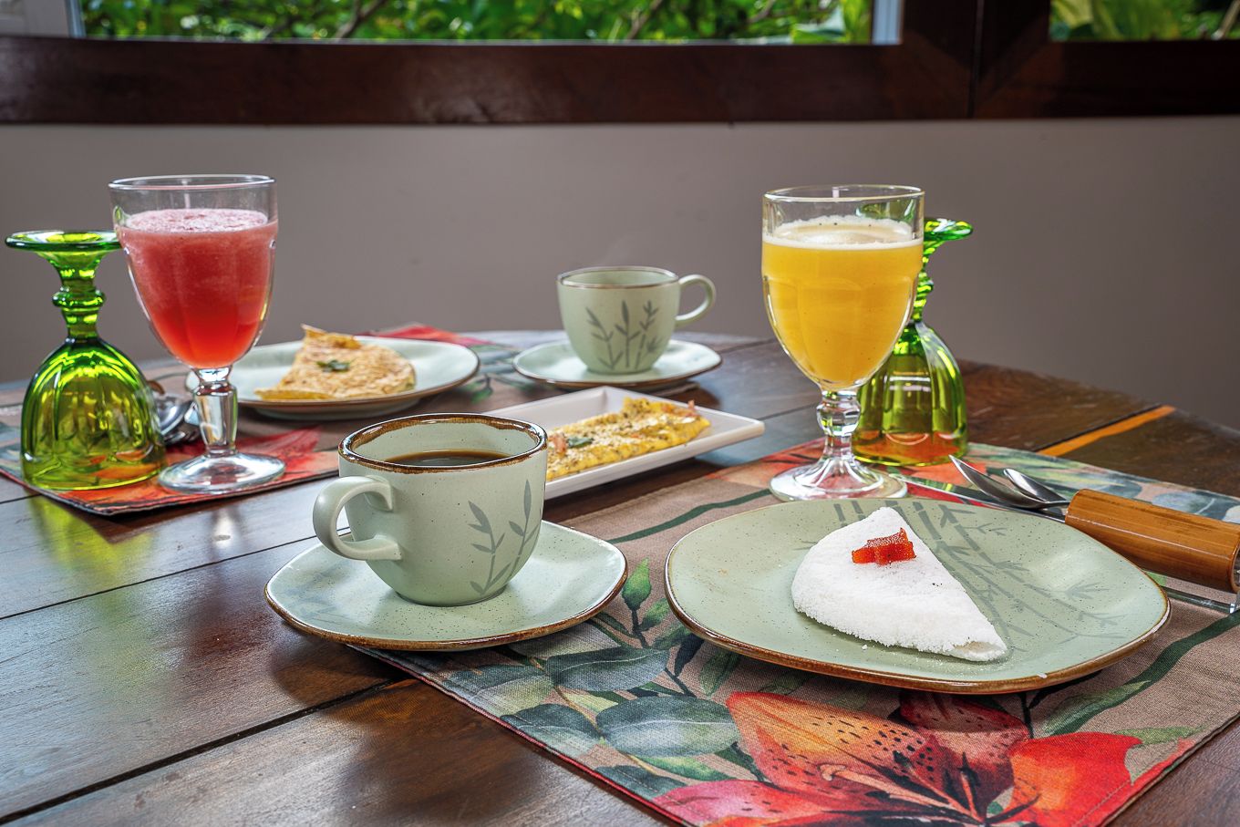 Ein Tisch gedeckt mit einer Tasse Kaffee, einem Glas Orangensaft, einem Glas rotem Getränk, einem Teller mit weißem Dessert und roten Früchten sowie einer grünen Flasche.