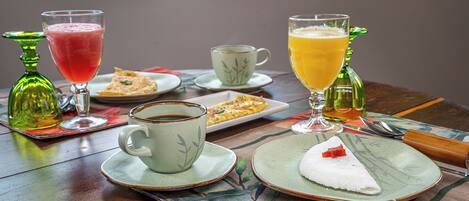 Ein Tisch gedeckt mit einer Tasse Kaffee, einem Glas Orangensaft, einem Glas rotem Getränk, einem Teller mit weißem Dessert und roten Früchten sowie einer grünen Flasche.