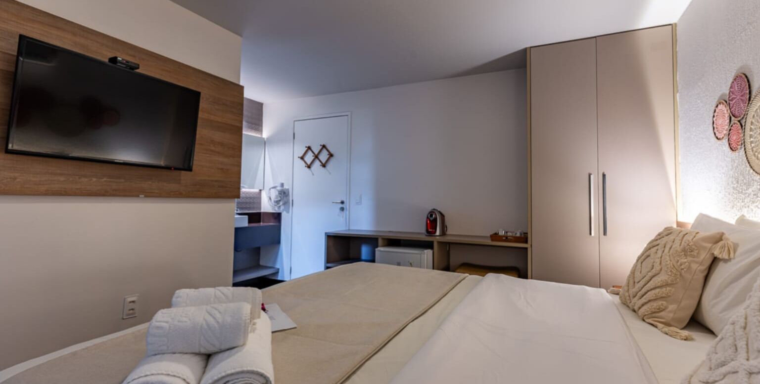A hotel room with a bed, a TV mounted on the wall, a wardrobe, and a desk with a kettle.