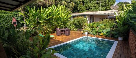 Un área de piscina con terraza de madera, rodeada de exuberante follaje y una casa al fondo.