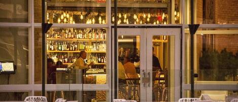 Un bar con scaffali pieni di bottiglie, un barista e sedie in vimini bianco davanti.