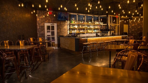 Eine Bar mit Theke, Hockstühlen und Tischen. An der Decke befinden sich Leuchten.