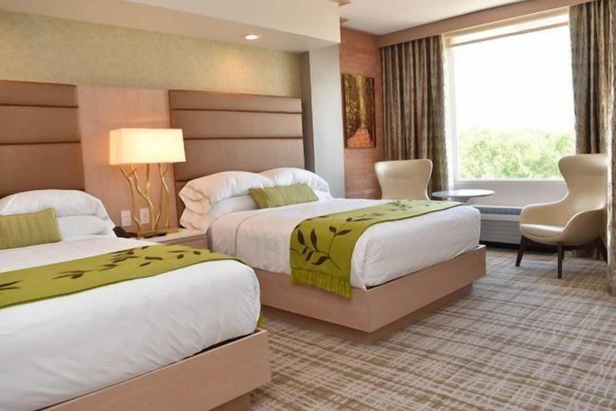 A hotel room with two beds, a nightstand, a lamp, a chair, and a window with curtains.