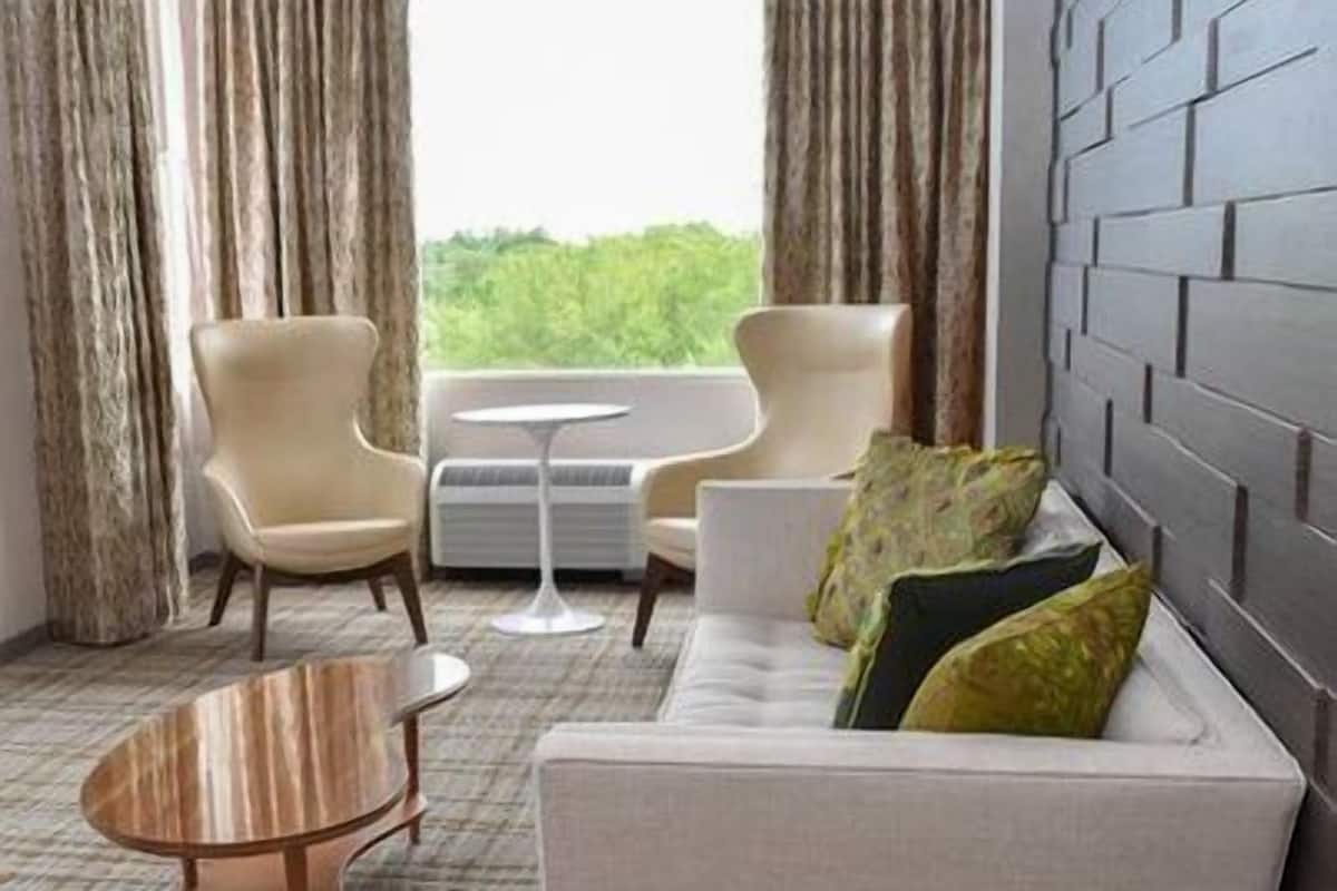 A modern hotel room with a sofa, two armchairs, a coffee table, and a view of greenery through the window.