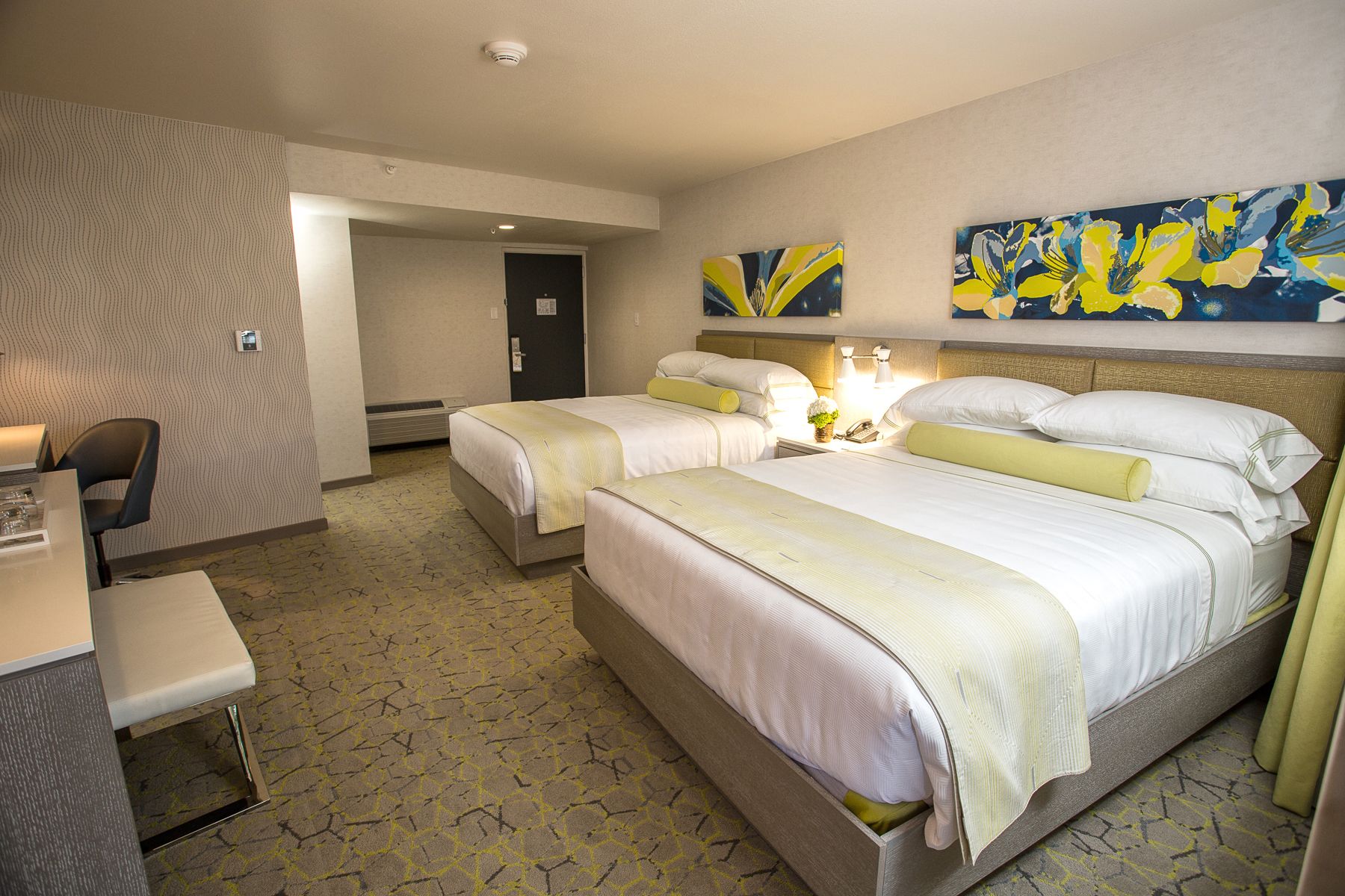 Une chambre d’hôtel avec deux lits, un bureau et une chaise. Une porte donne accès à une autre pièce, et un mur est décoré d’œuvres d’art abstraites.