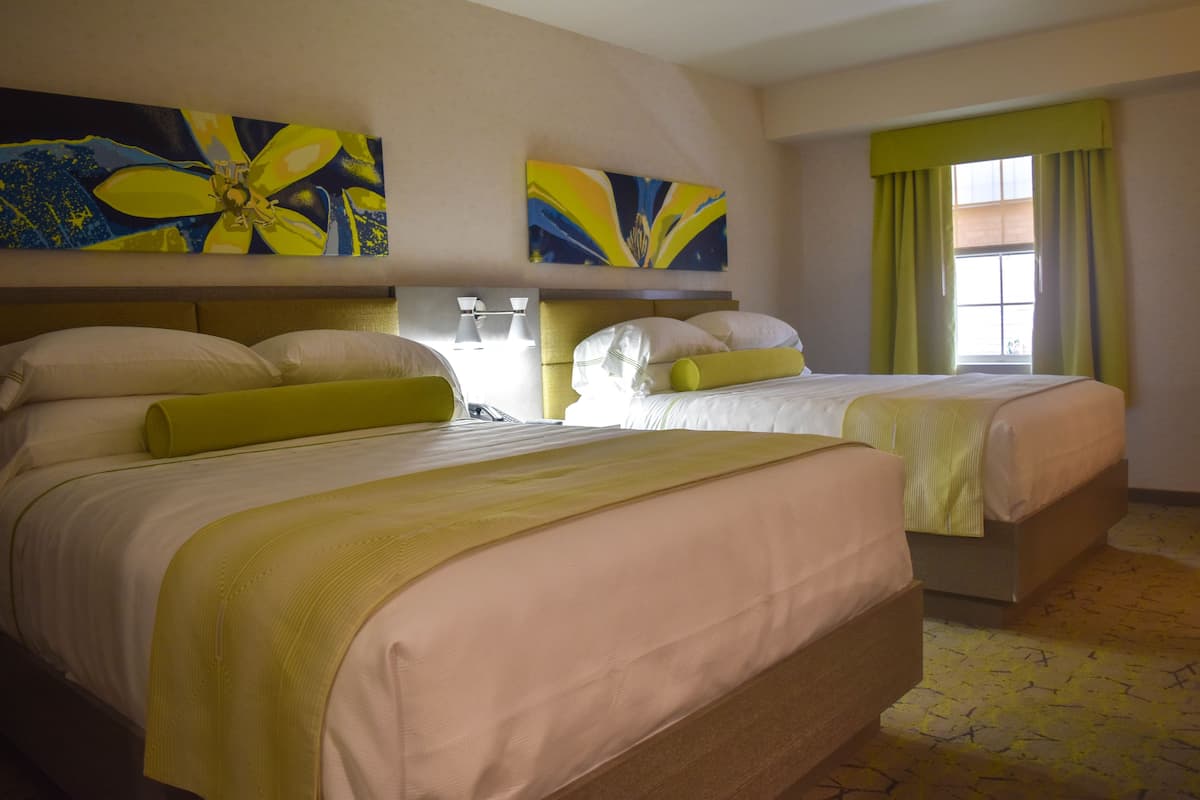 A hotel room with two beds, each with a headboard, a large window with curtains, and two abstract paintings on the wall.