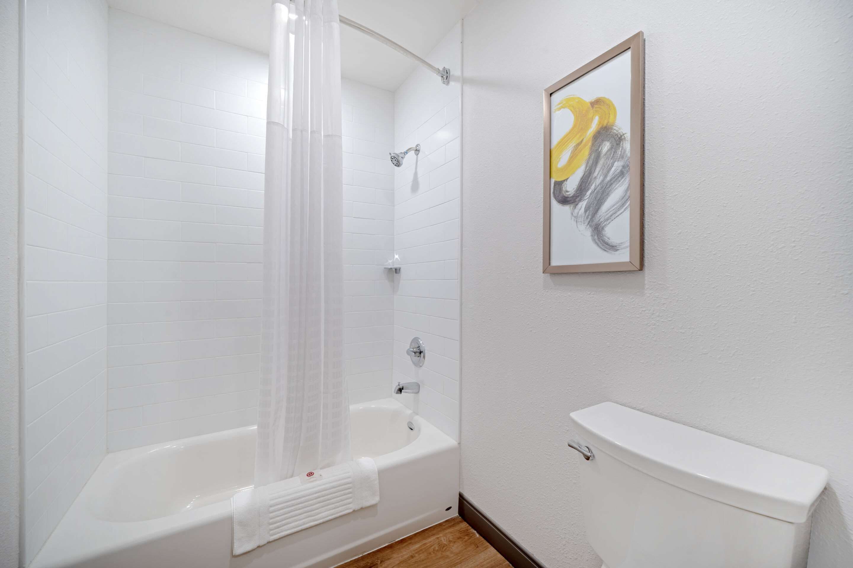 A bathroom with a white bathtub, a shower curtain, a toilet, and a framed artwork on the wall.