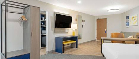 A hotel room with a bed, desk, chair, TV, wardrobe, and a small seating area.