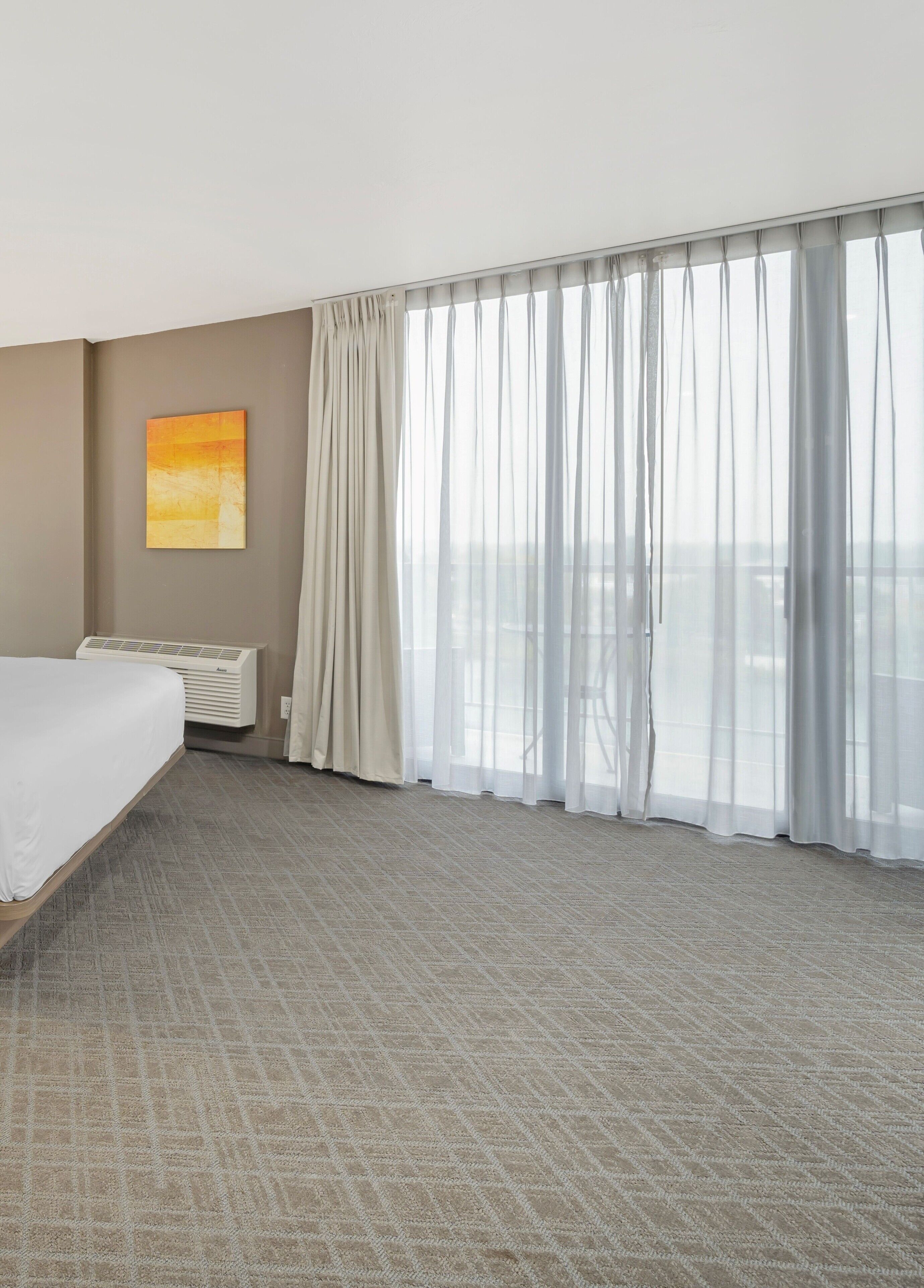 Una habitación de hotel con una cama grande, aire acondicionado montado en la pared, una ventana con cortinas translúcidas y un cuadro en la pared.