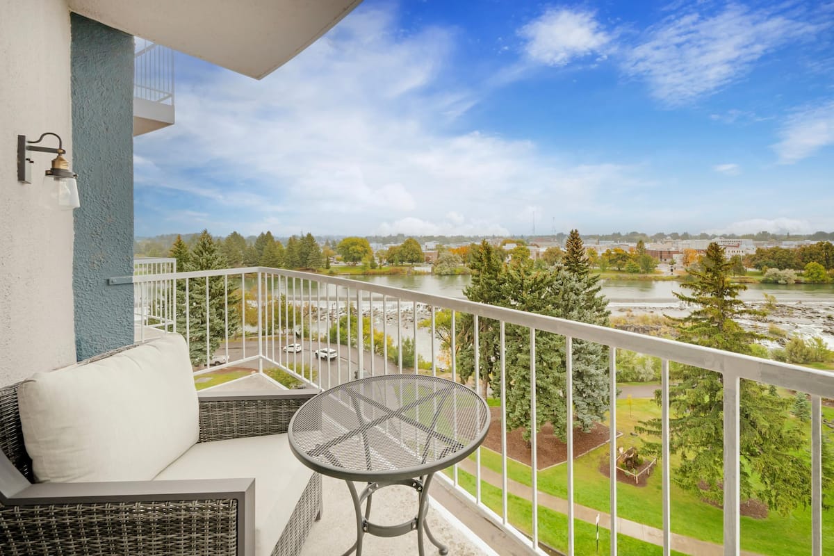 Ein Balkon mit einer Rattan-Couch, einem runden Metalltisch und Blick auf einen Fluss und einen Park.