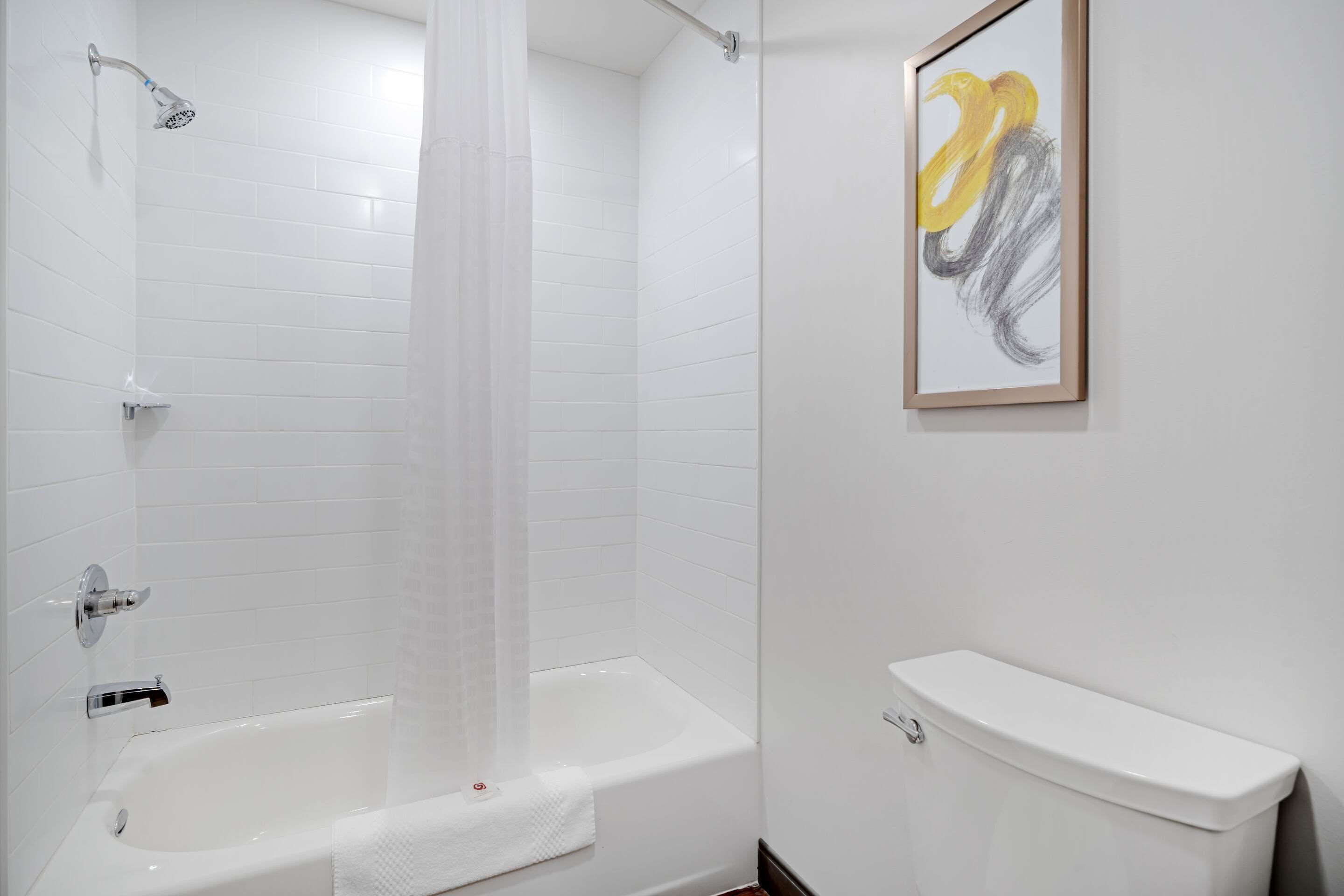 A bathroom with a white bathtub, a showerhead, a toilet, and a framed abstract painting on the wall.