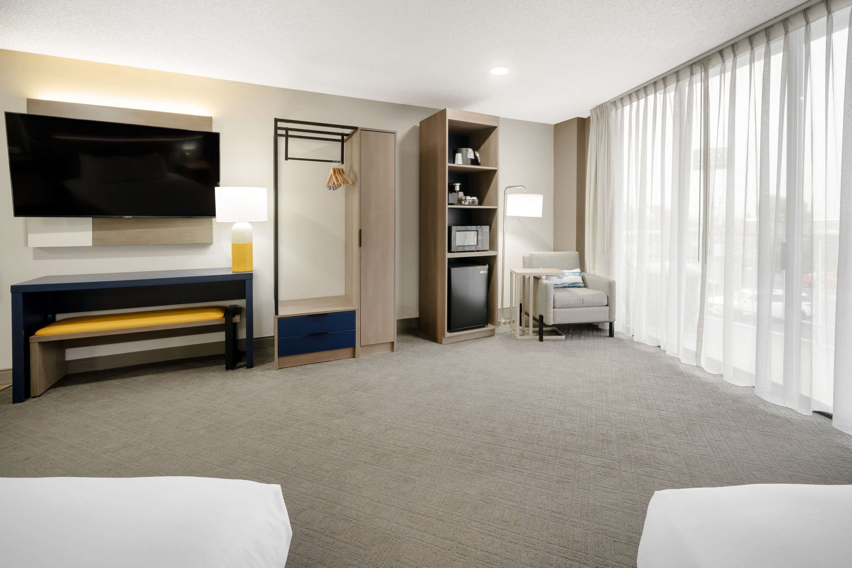 A hotel room with a large flat-screen TV, a desk with a bench, a sofa, a small table with a lamp, and a large window with sheer curtains.