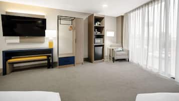 Hotelkamer met een groot flatscreen televisietoestel, een bureau met een bankje, een sofa, een kleine tafel met een lamp en een groot raam met dunne gordijnen.