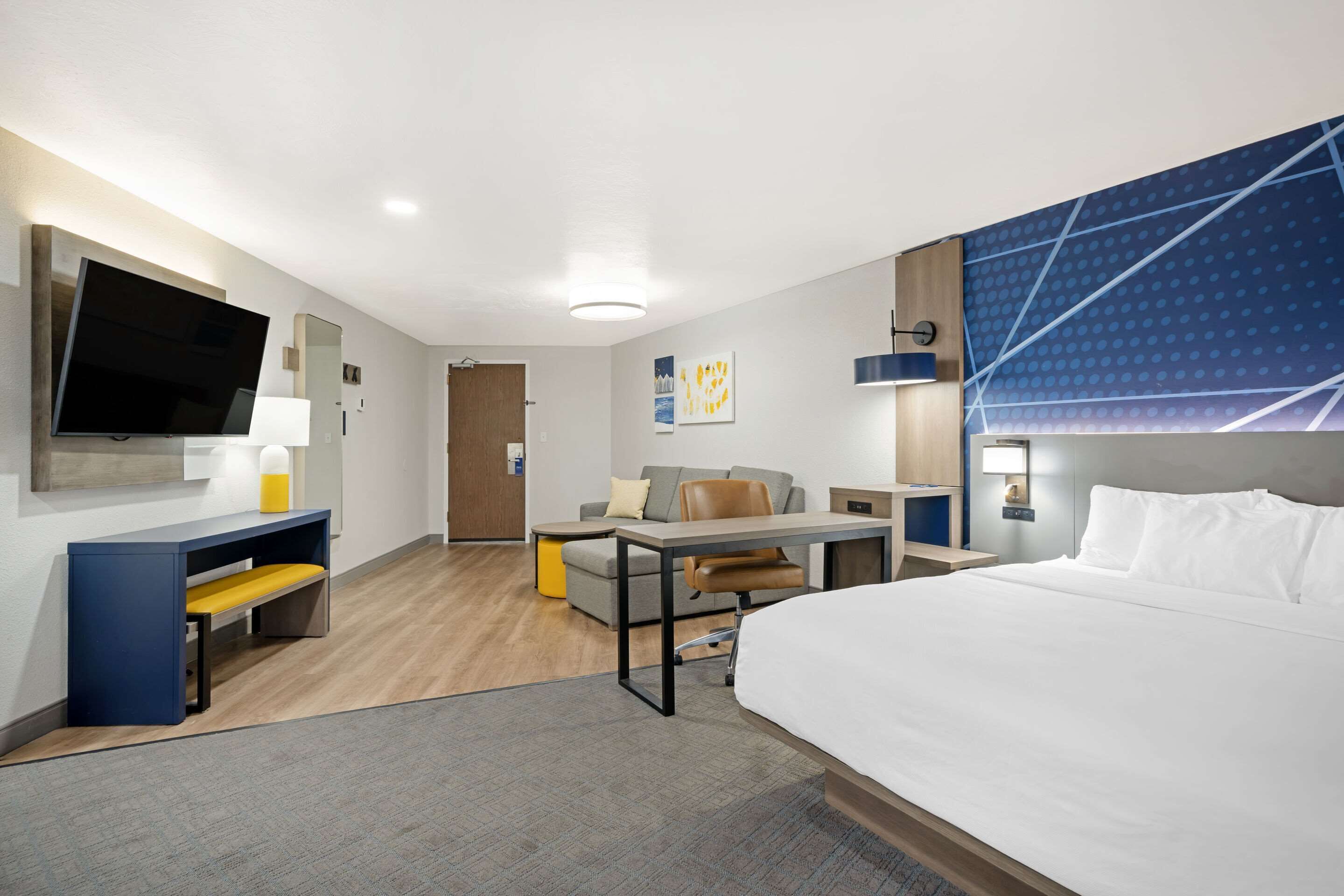 A modern hotel room with a large bed, a desk with a chair, a sofa, and a TV.