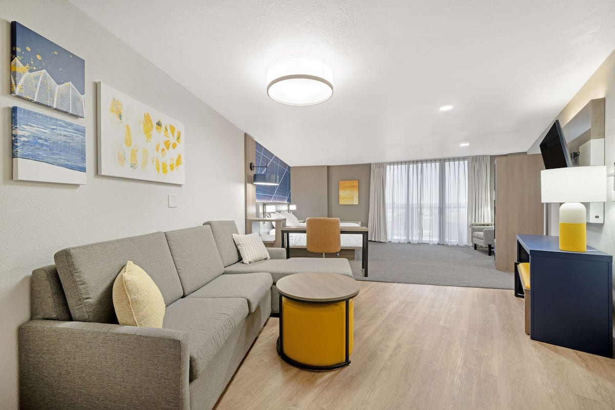 Ein modernes Hotelzimmer mit einer Eckcouch, einem runden, gelben Hocker, einem Essbereich mit Tisch und Stühlen sowie Bildern an den Wänden.