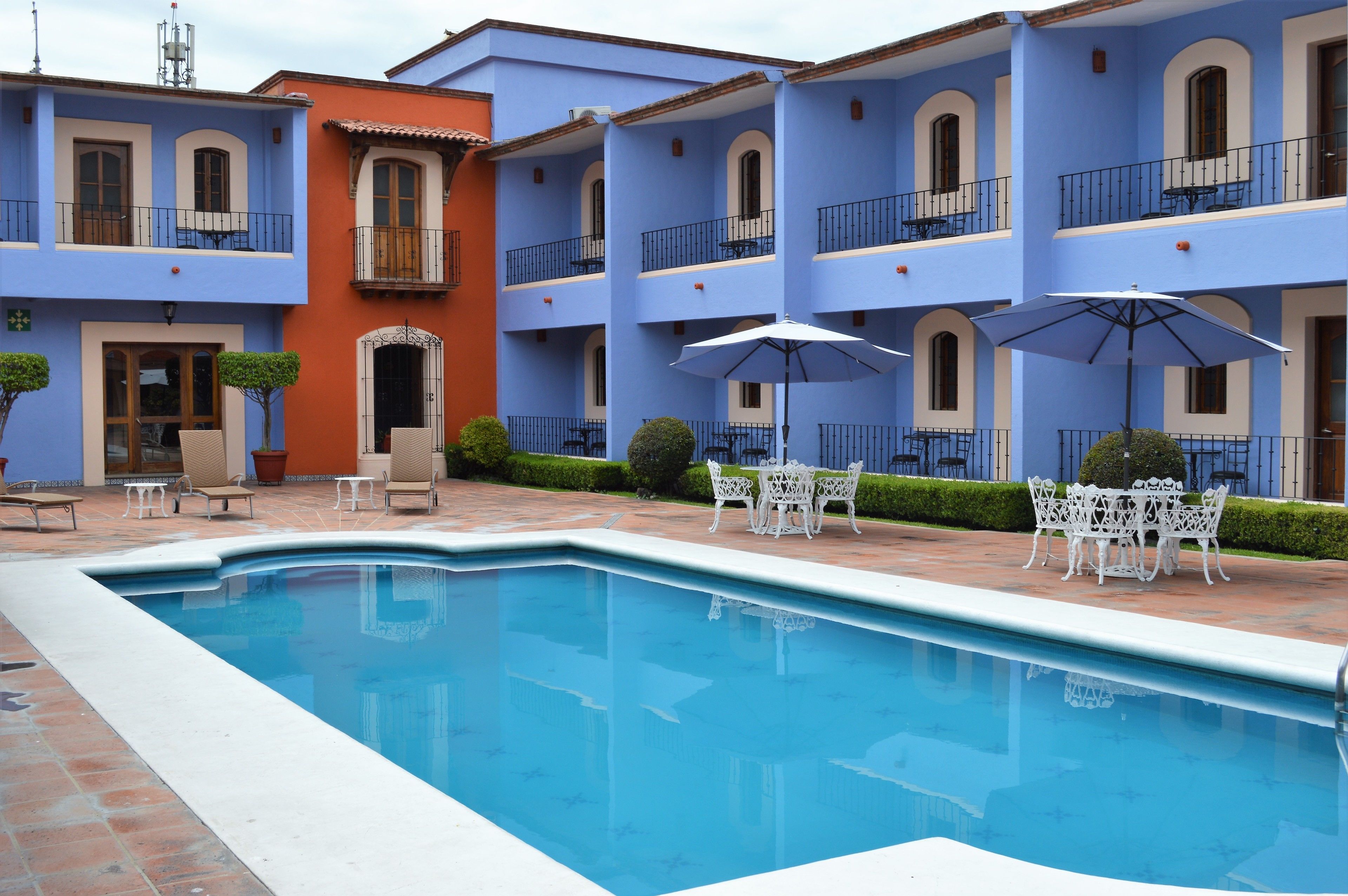 Una piscina con mobiliario blanco de exterior y sombrillas, rodeada por un edificio con fachadas azules y anaranjadas.