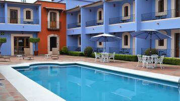 Una piscina con mobiliario blanco de exterior y sombrillas, rodeada por un edificio con fachadas azules y anaranjadas.