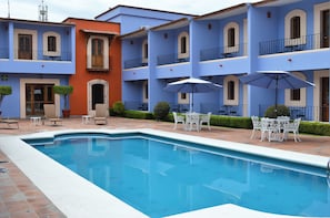 A swimming pool with white outdoor furniture and umbrellas, surrounded by a building with blue and orange facades. - Hotel Santa Helena Plaza (Oaxaca)
