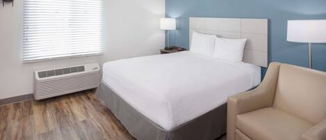 A hotel room with a large bed, a bedside table with a lamp, a chair, and a window with blinds.