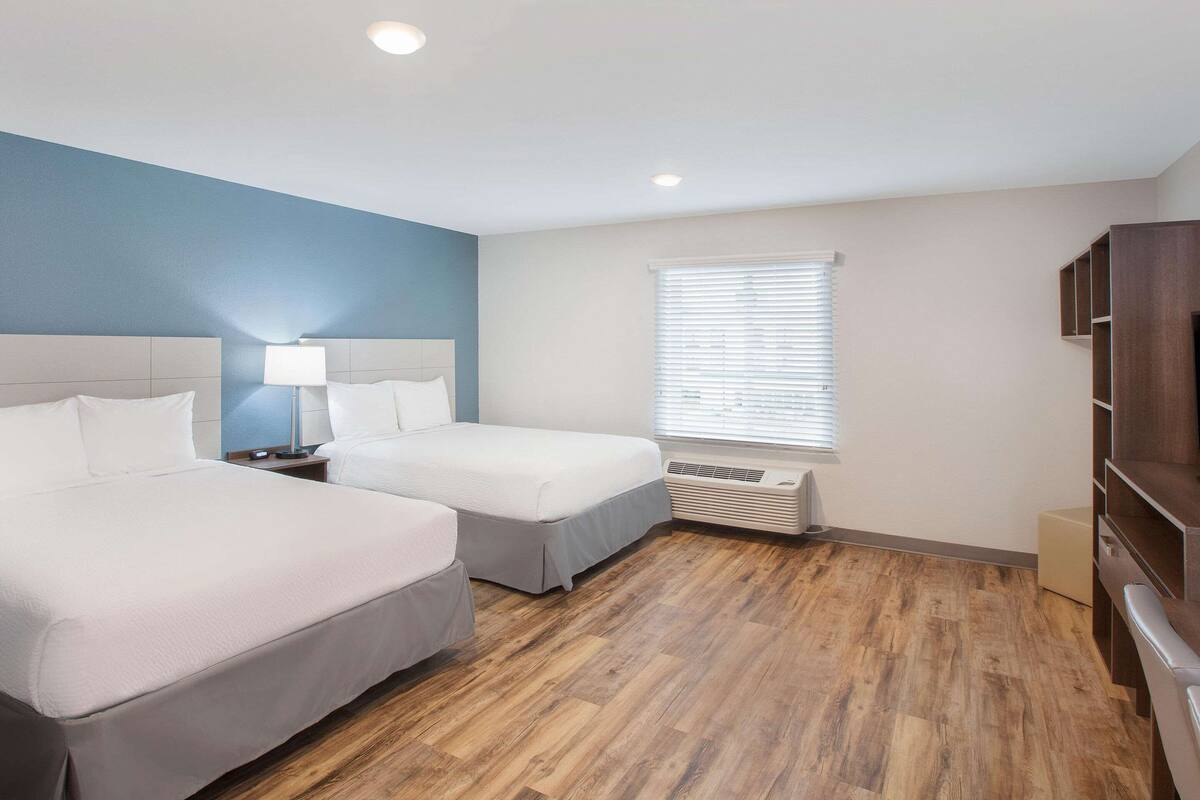 Quarto de hotel com duas camas, uma mesa de cabeceira com abajur, janela com persianas e prateleira embutida para televisão.
