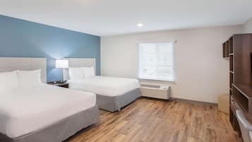 A hotel room with two beds, a nightstand with a lamp, a window with blinds, and a built-in TV shelf.
