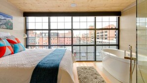 A bedroom with a large window, a bed with colorful pillows, a blue throw, a white bathtub, and a city view.