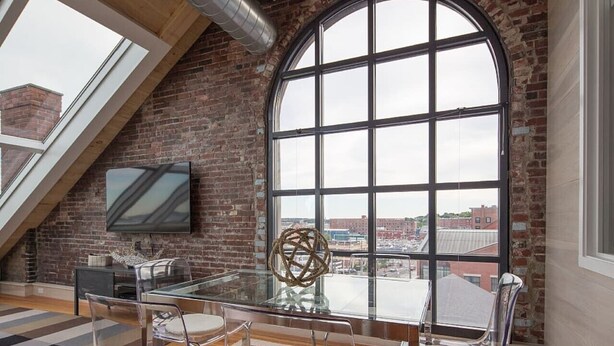 A modern loft with a large arched window, a glass dining table, and a flat-screen TV mounted on the brick wall.