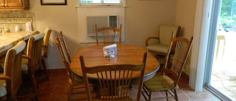 Comedor con una mesa redonda de madera rodeada de sillas de madera, una ventana con persianas y una zona de cocina con armarios de madera.