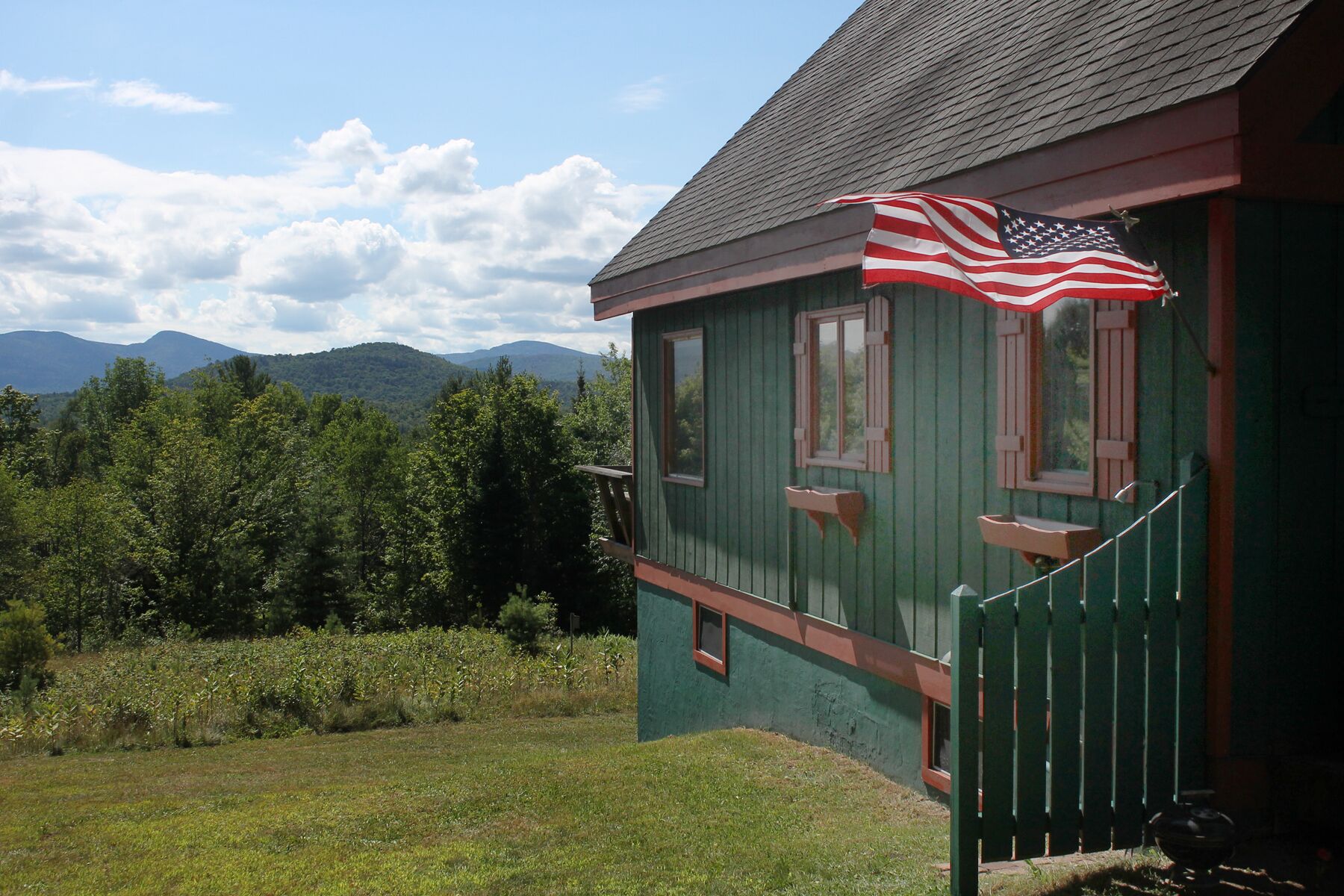 A green wooden house with a porch, an American flag, and a view of mountains.