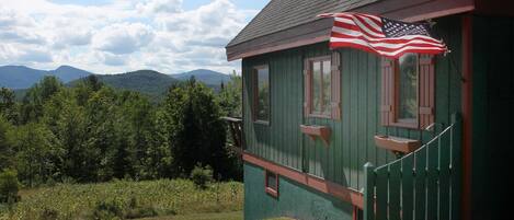 A green wooden house with a porch, an American flag, and a view of mountains.
