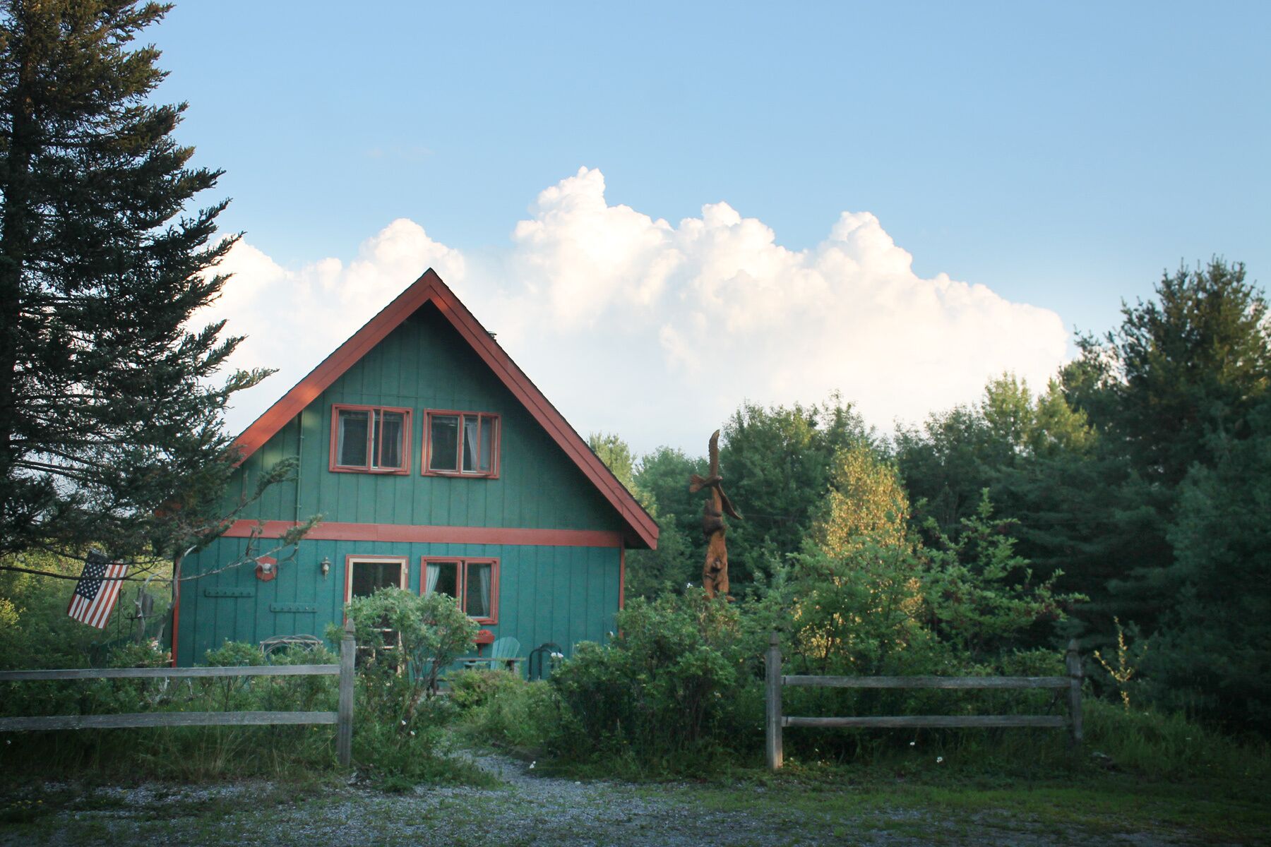 Une maison verte avec un toit rouge, entourée d’arbres et d’une clôture en bois.
