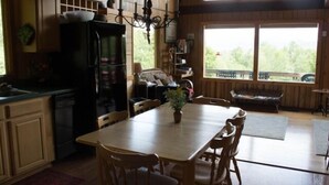 A wooden interior with a dining table, chairs, a chandelier, and a large window with a view of greenery. - Mountain View Chalet (Indian Lake)