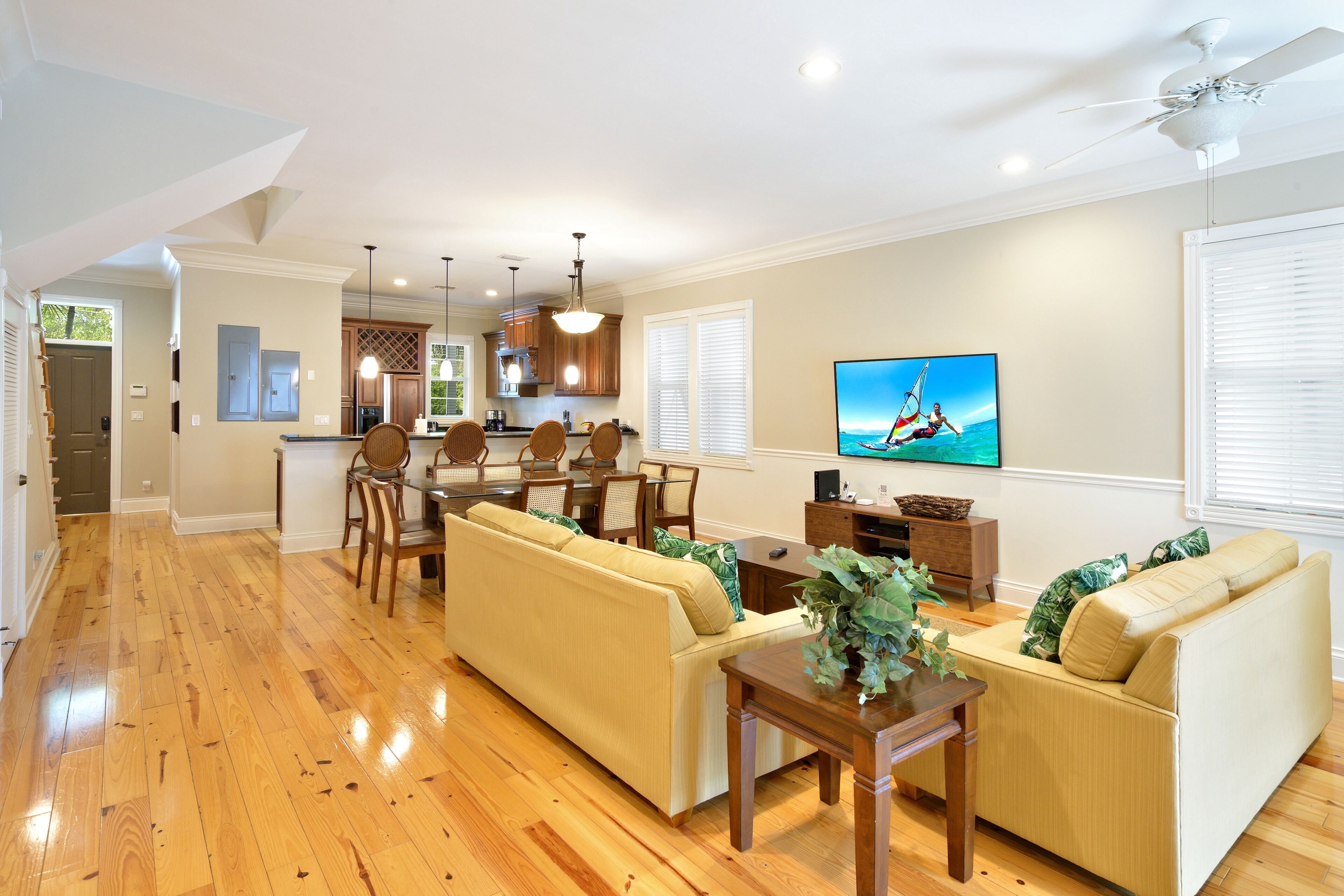A spacious living room with a wooden floor, a beige sofa set, a dining area with chairs and a table, a kitchen in the background, and a TV mounted on the wall.