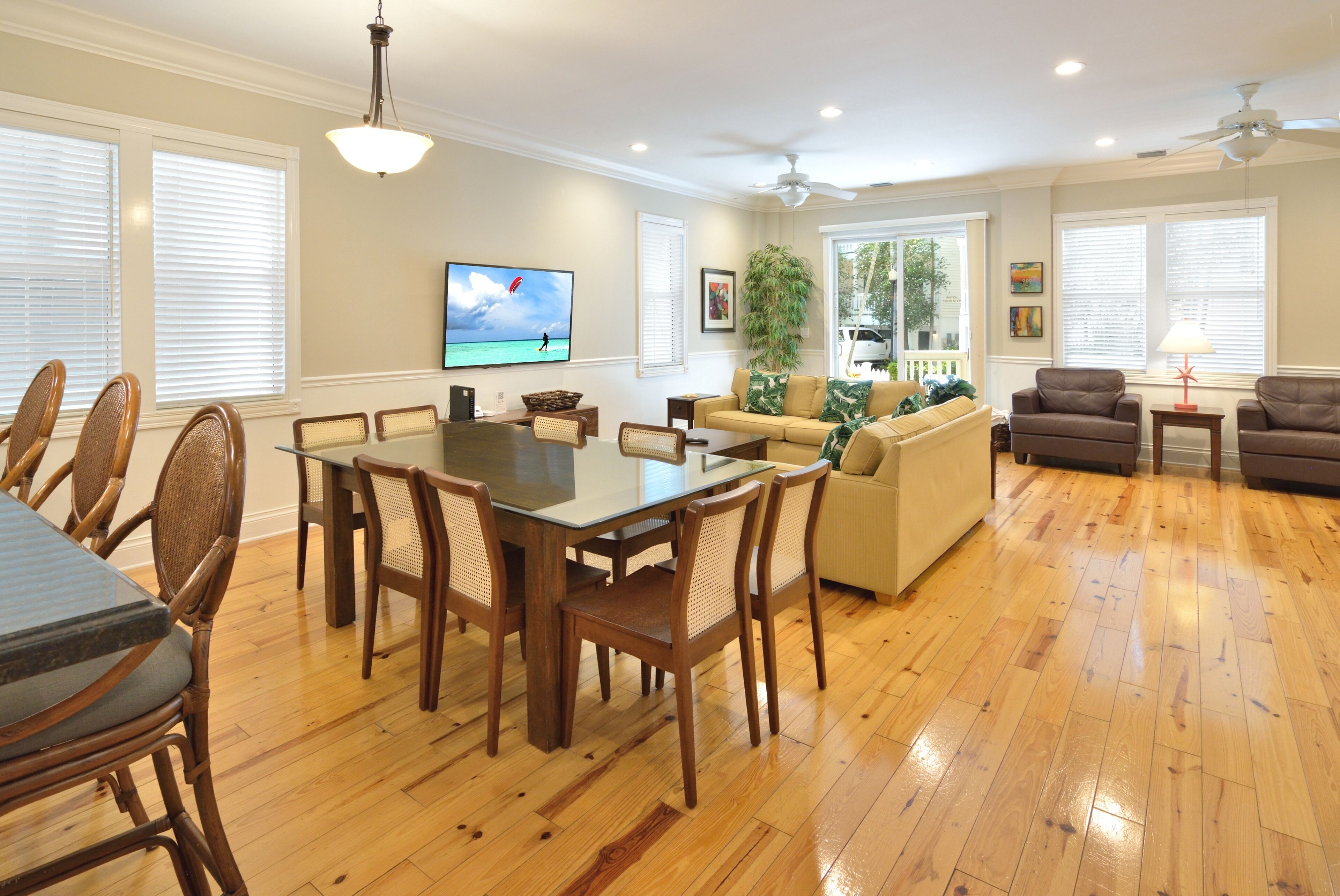 A spacious living room with a dining area, wooden flooring, and a large TV displaying a beach scene.