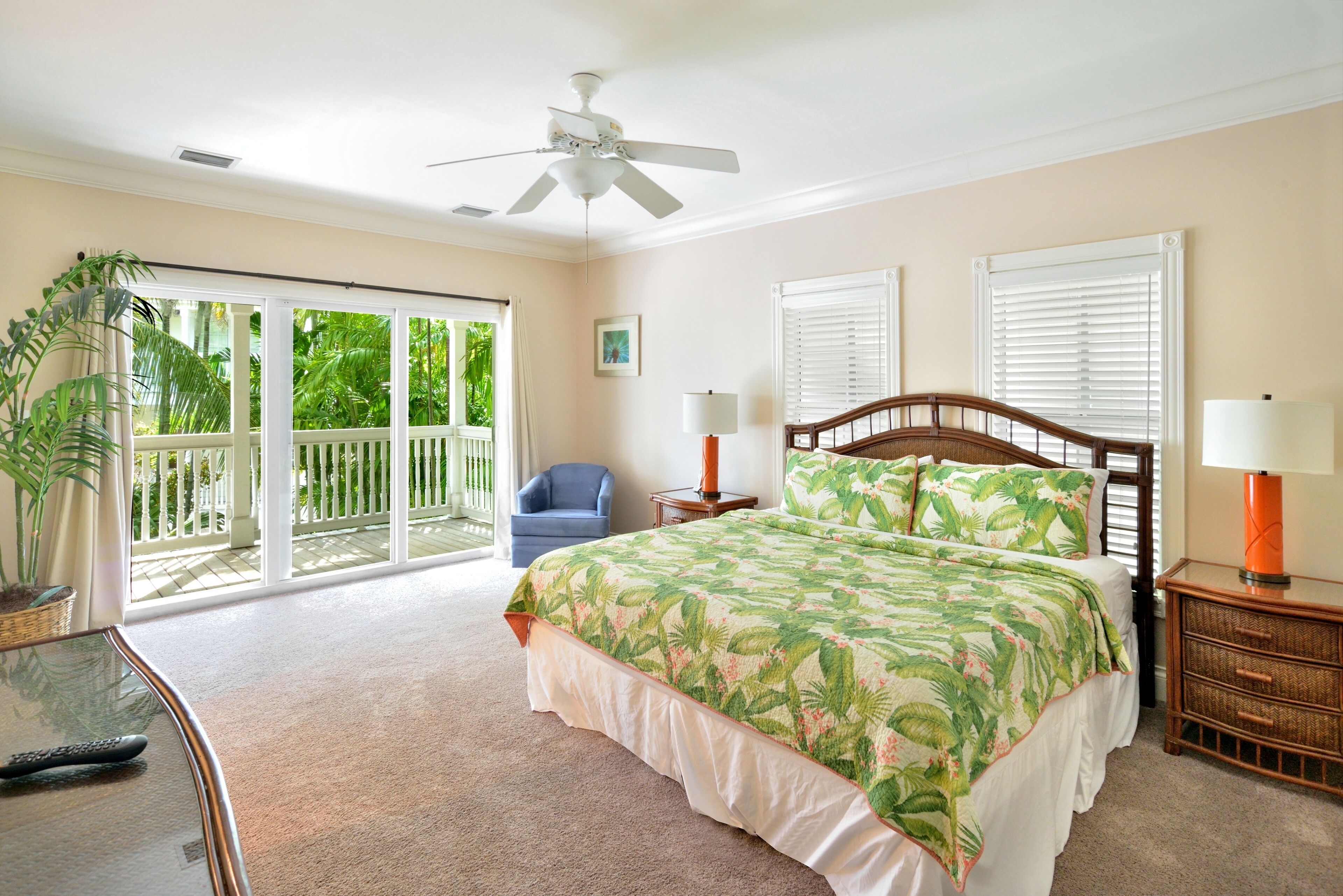 A bedroom with a bed, a ceiling fan, a nightstand, a lamp, a chair, and a balcony with a view of greenery.