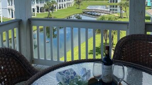 Un espace extérieur abrité, avec une table dressée pour deux personnes, agrémenté d’une bouteille de vin et de verres, donnant sur une zone verdoyante avec un plan d’eau et des bâtiments en arrière-plan.